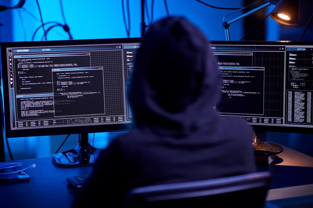 Hacker in a dark room, wearing a hoodie, working on multiple computer screens with code.