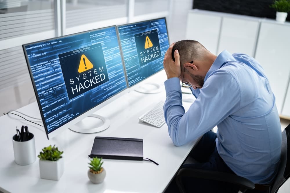 A person sits at a desk with two monitors displaying "SYSTEM HACKED" error messages.