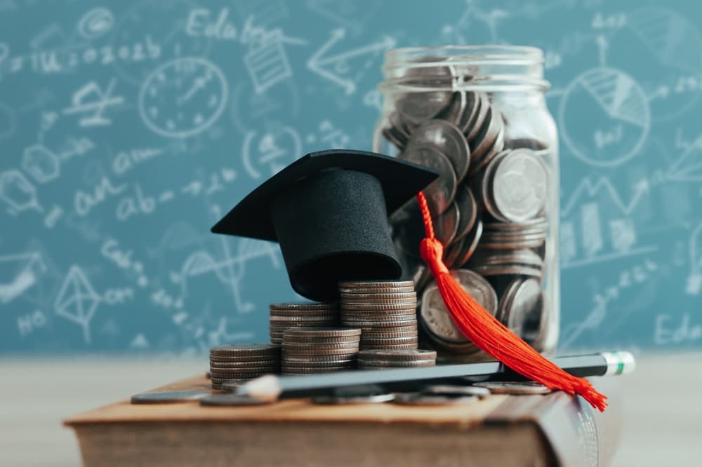 A graduation cap on stacks of coins next to a jar filled with coins and a book.