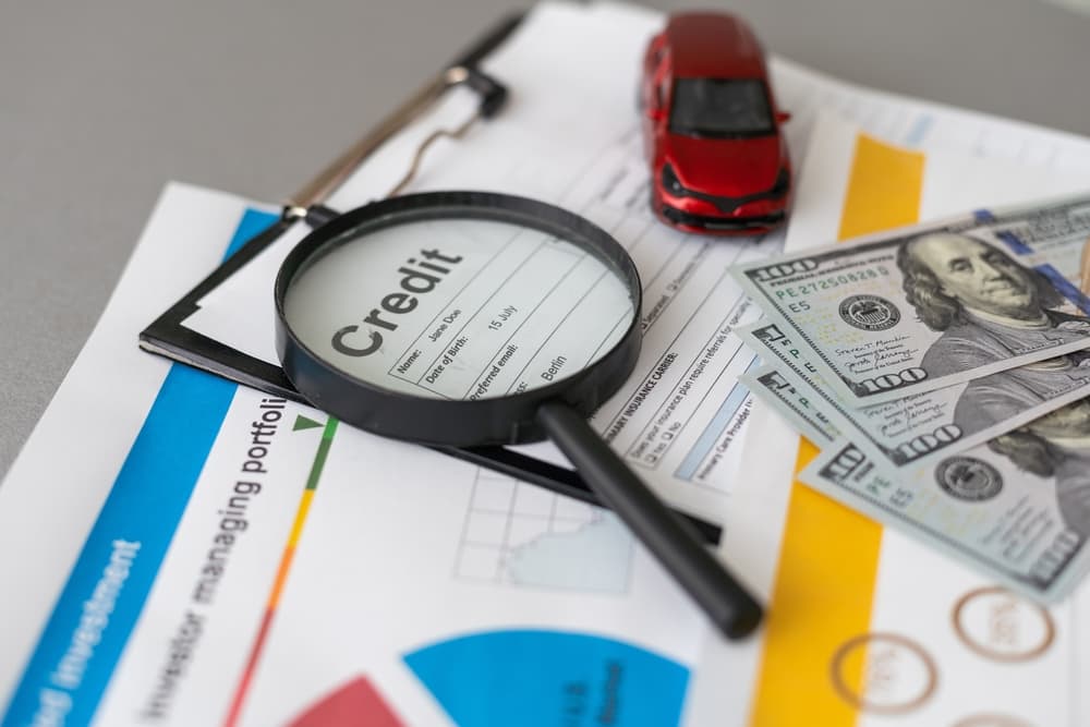 Magnifying glass focusing on a credit report with cash, documents, and a toy car nearby.