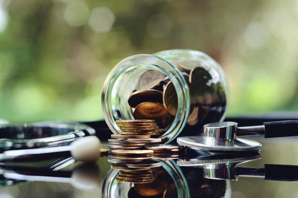 Coins spilling from a jar beside a stethoscope on a reflective surface.