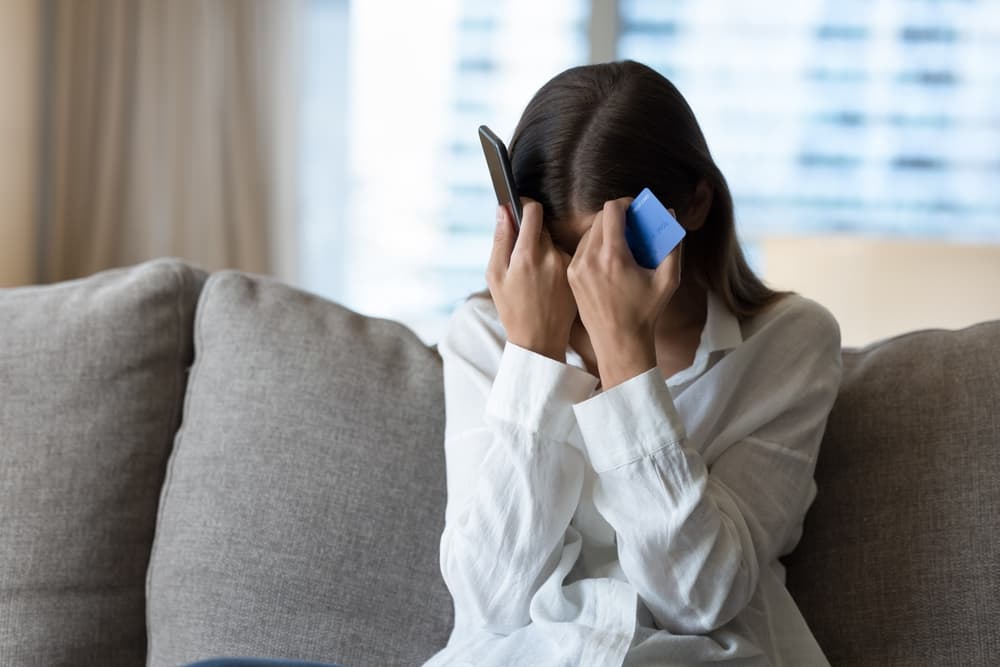 Distressed woman holding phone and credit card, sitting on a couch, overwhelmed.