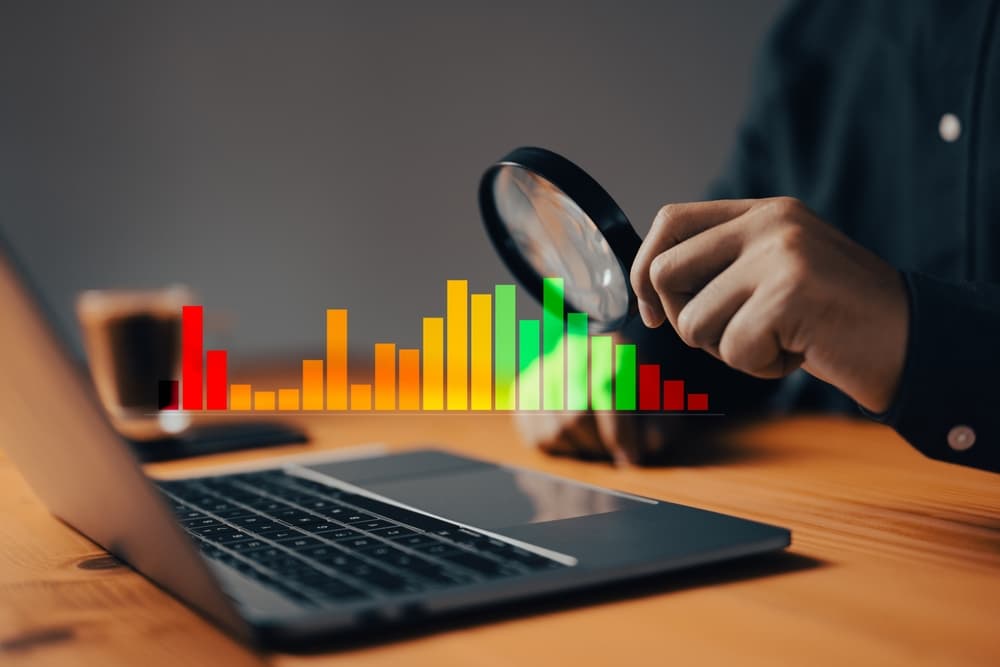 Person using magnifying glass to examine colorful bar graph on a laptop screen.