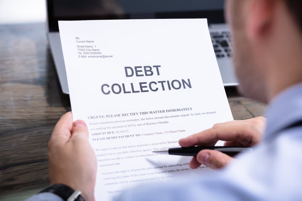 Person reading a debt collection notice, holding a pen, with a laptop nearby.