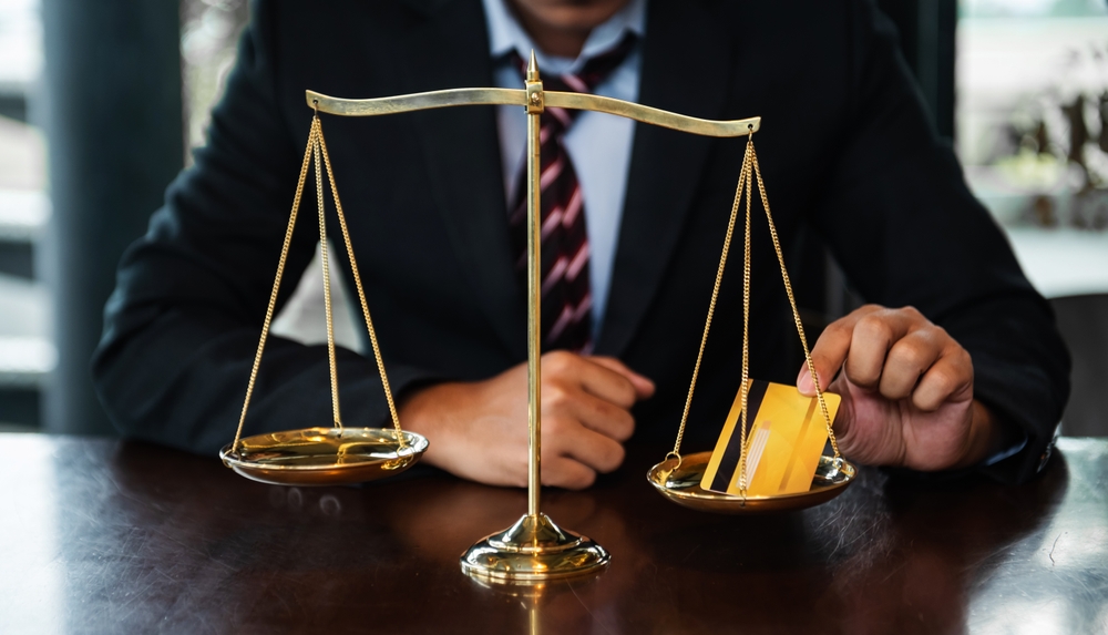 Close-up of a lawyer's hand holding a credit card, with money and a scale of justice in a courtroom, symbolizing a credit card agreement.