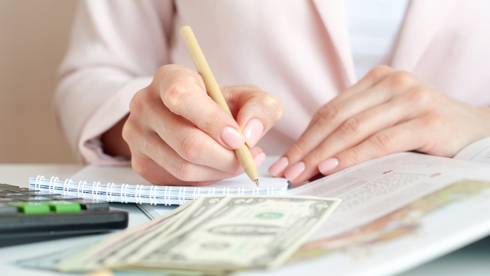 Close-up of a woman using a calculator and notebook, with a pink background, managing finances through debt consolidation.