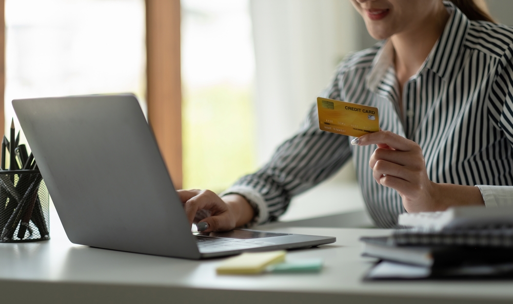 Asian woman with laptop and credit card at home, smiling as she learns about the basics of credit card agreements.