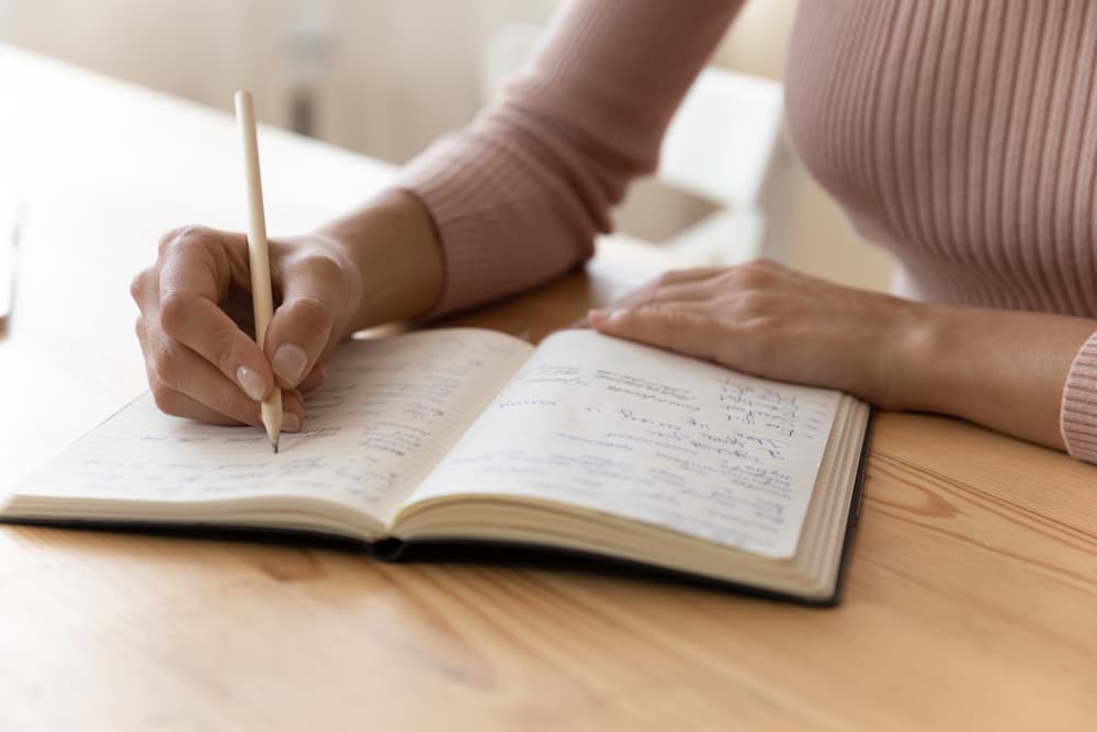 Millennial woman writing notes in a notebook at a desk, focusing on work or study tasks at home.