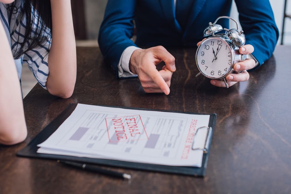 Close-up of a debt collector holding an alarm clock, pointing at foreclosure and final notice documents on a table near a woman.