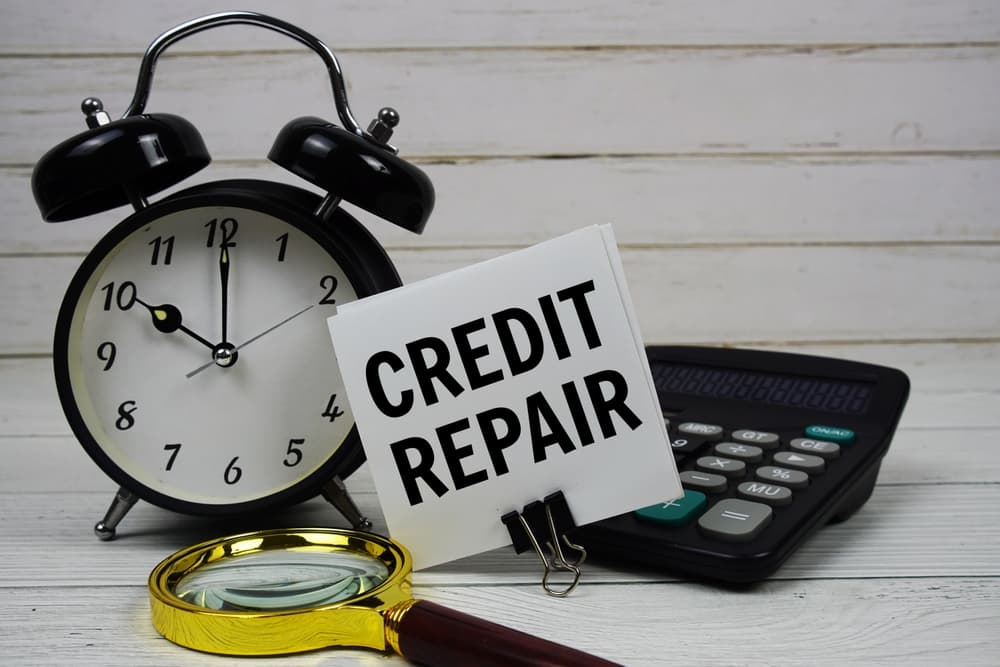 Credit Repair written on a paper card next to an alarm clock, calculator, and magnifying glass on a wooden background.